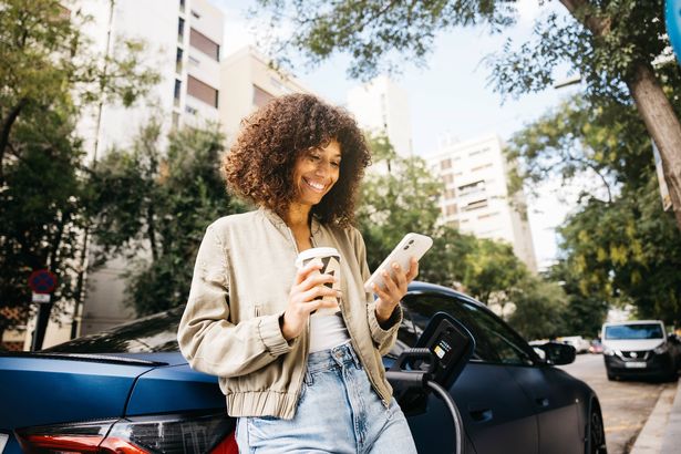 A woman enjoys her coffee and phone while her electric car charges in an urban setting.