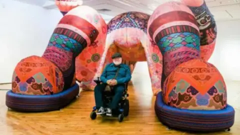 Hull City Council Artist Jason Wilsher-Mills sat in a wheelchair wearing a blue top, flat cap and blue trousers and trainers, with one of his large interactive sculptures filled with different colours.