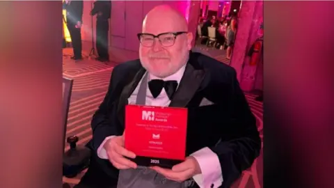 Ferens/Hull City Council Jason Wilsher-Mills holding his award and smiling at the camera, wearing a bow tie and a suit
