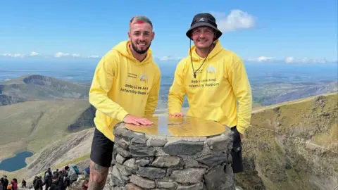 Joel Redhead Joel Redhead with multi-colour hair and a black beard stands next to his friend Shane who is wearing a blue hat on the summit of Snowdon. The pair are wearing yellow hooded tops with the Chasing Rainbows charity logo on them.