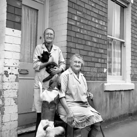 Alec Gill A black and white photograph of two women, one of them elderly, standing outside a brick house. One is holding a cat, smiling, while the other sits with a cane, also smiling. A dog jumps up toward the cat.