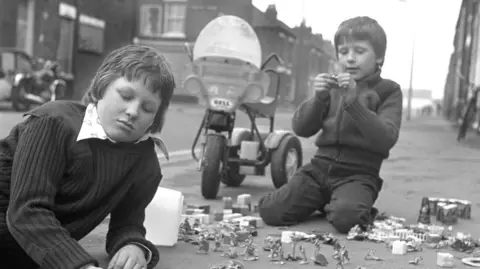 Alec Gill A black and white photograph of two boys playing on a street, kneeling on the pavement surrounded by toy soldiers and miniature buildings. One boy is focused on a small motorcycle toy. A tricycle with a windshield is behind them.