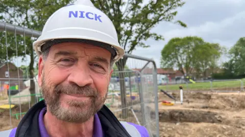 BBC TV presenter Nick Knowles smiles into the camera. He has a beard and is wearing a white hard hat, purple t-shirt, black top and a purple high-vis vest. He is standing in front of a building site and a metal fence. Trees and houses are visible in the distance.