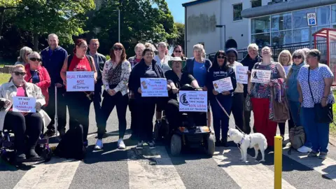 Natalie Bell/ BBC A group of men and women, the majority middle-aged or elderly, stand in the grounds of a leisure centre building in bright sunshine. One woman is sitting on a wheelchair, another in a mobility scooter. One holds a white dog on a lead. Some have placards with slogans reading: "Real trainers, not AI screens", and "Would you take 40% less income."