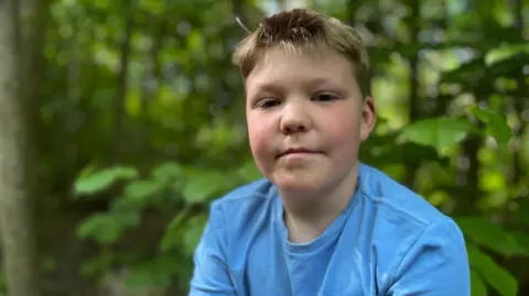 Jo Makel /BBC A boy, in woodland is pictured looking at the camera. He has blonde hair and is wearing a blue t-shirt