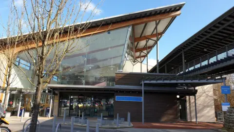 HULL UNIVERSITY TEACHING HOSPITALS NHS TRUST Glass building with sloped roof. A smaller reception structure in-front and to the side. There are trees and bollards with paving.