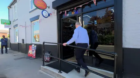 BBC A man wearing a blue shirt and dark trousers walks up a concrete wheelchair-accessible ramp to the large black-framed glass doorway of a village shop. The brick building is painted light-grey and black. Signs advertise a Post Office, the National Lottery and ice creams. A man stands to the left wearing a blue jacket.