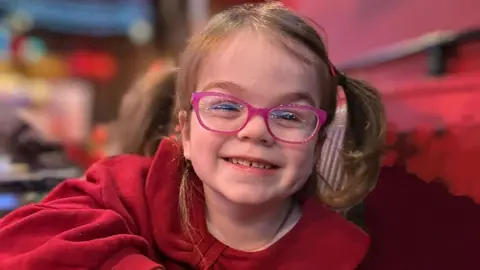 Family Tilly smiling at the camera she has brown hair in ponytails and is wearing pink glasses and a red dress