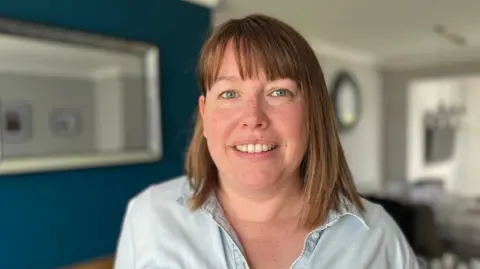 BBC/Anne-Marie Tasker A woman with brown shoulder-length hair, smiling at the camera. She is wearing a pale blouse. There are blue and white walls behind her with mirrors on them.