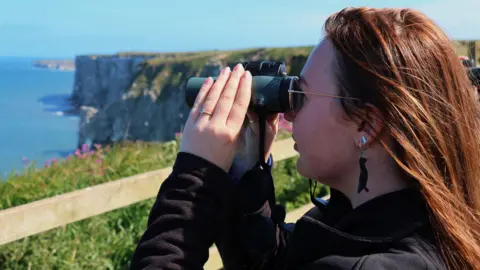 Ellen Leach A woman stands on the cliffs at Bempton, East Yorkshire and looks out to sea using a pair of black binoculars. She has long red hair and is wearing a black jacket. The side of her face is visible and she has an earring in the shape of a bird. In the background, a wooden fence and exposed cliff is visible along with the sea on the left of the image.