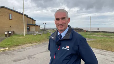 BBC / Simon Spark RNLI operations manager David Scott. He is wearing a dark blue jacket with RNLI Lifeboats emblazoned on it. He is standing outside the RNLI station with the sea in the background. 