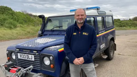 BBC / Simon Spark RNLI regional estates manager Gary Aarnes-Olsen. He is wearing a dark blue RNLI-branded fleece. He is standing in front of an RNLI land rover Defender. 
