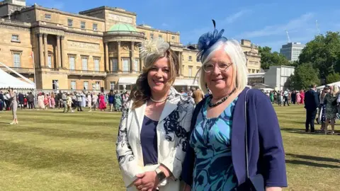 Emily Griffin Two ladies are smiling at the camera. They are dressed smartly with fascinators on - the lady on the left in white with a white headpiece, and the lady on the right in blue with a blue headpiece. They are stood in front of a grand building, and in the background there are lots of people stood on the grass.