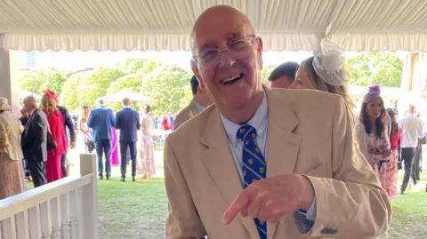 Roger Vine A man is seen smiling directly at the camera. He is wearing a beige suit with a blue striped tie. He has silver thin glasses and is seen to be pointed at something off camera.