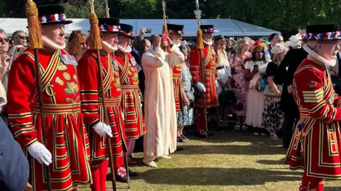 Emily Griffin Six beefeaters are seen amongst a crowd of people all smartly dressed. They are in bright red and gold clothing, with staffs adorned with a gold tassel in their hands. They wear black hats, with red, white and blue flowers around the rim. A man with a pink hat and a cream dress is seen in the middle.