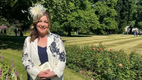 Emily Griffin A lady is smiling at the camera. She is smartly dressed in a cream blazer with navy flowering detailing, and also has a matching cream fascinator on her head. She is stood in front of lush greenery of trees, hedges and a well-manicured lawn.