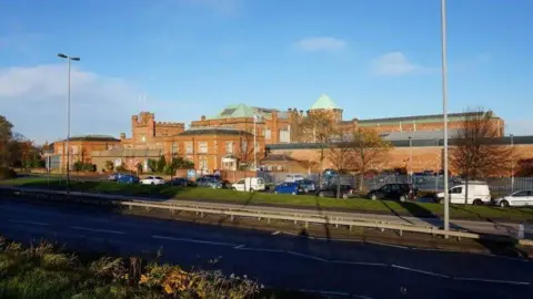 Geograph/Ian S Brick building with green roof. Some of the roof is pointed and other parts are like a castle's battlements. There are cars parked in front and a road. There are bushes and grass.