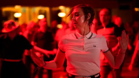 Getty Images A woman with short brown hair and wearing a white polo shirt dances to Northern Soul music. She is bathed in red and white light and other dancers are visible in the background.