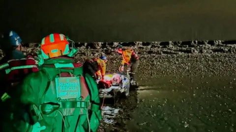 Humber Rescue The Humber rescue team attend a casualty, who cannot be seen, on the the edge of the estuary. The team wear helmets and hi-vis suits with "Humber Rescue" written on them. The scene is illuminated at night, revealing a muddy shore and dark water.