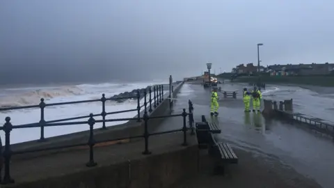 Jim Blake Sea wall and metal rails and rough sea on one side and three people in fluorescent stood on a wet promenade.