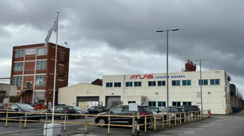 Richard Madden/BBC The exterior of Atlas Leisure Homes' headquarters. The main building is covered in cream cladding and has a large blue and white logo spanning the width of the building. A building on the left is a red brick tower block with six large windows. A car park is in front of the two buildings, as are a flagpole and lights.