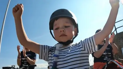 BBC Henry sat on his bike with hands aloft after completing his race he wears a helmet, a ventilator tube and a striped blue and white T-Shirt. behind him other people on bikes are applauding him.