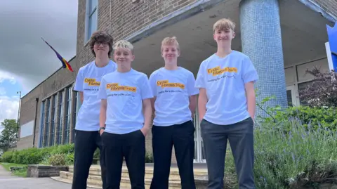 Family handout Four boys. Three have short blonde hair and one has slightly longer brown hair. They are smiling. Three have dark trousers and one has grey trousers. They are wearing white t-shirts with Cystic Fibrosis Trust written on them. They are stood in front of a brick building, a flag, tarmac and plants.