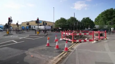 BBC The junction of Princes Avenue and Spring Bank West in Hull. The road, which features multiple sets of traffic lights, is covered with orange and silver cones. A red and white barrier surrounds a large hole and a large pile of rubble. An industrial building stands in the distance.