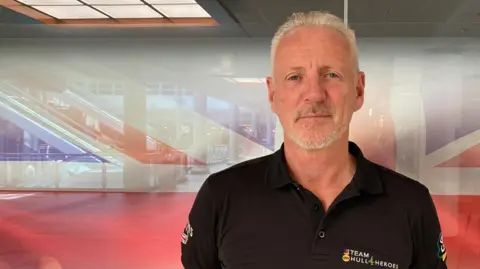 Sally Fairfax/BBC Head and shoulders shot of a man with grey hair and goatee beard standing in front of a Union flag. He is wearing a black polo shirt and has his arms behind his back.