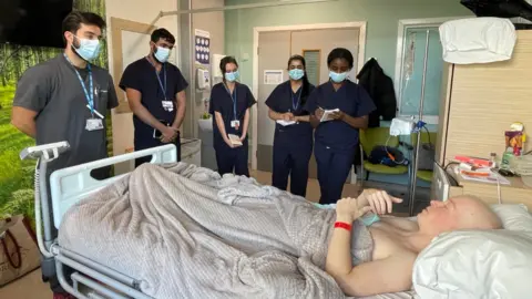Hull University Teaching Hospitals NHS Trust A man in a hospital bed having lost his hair through cancer treatment surrounded by five doctors at the bedside taking notes