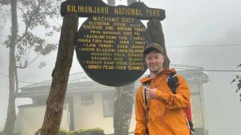 Hull University Teaching Hospitals NHS Trust Freddie wears an orange raincoat and points to a sign that reads Kilimanjaro national park, that also includes timings to different points within the park. It is very misty and wet.