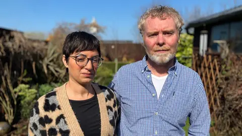 BBC Asha and Jason Ross are standing side by side outdoors. Asha is wearing a beige cardigan with black spots over a black shirt, and has short dark hair. Jason is wearing a blue and white checkered shirt over a white undershirt, and has curly light-colored hair. The background shows greenery, including bushes and trees, with a clear blue sky above. 