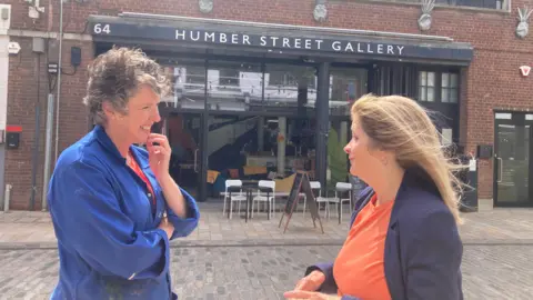 BBC / Sally Fairfax Lady smiling with short wavy hair, blue denim jacket and striped red and orange jumper with lady with long brown hair, blue jacket and orange top. They are standing on a paved street in front of a brick and glass building with tables and chairs outside of it and an advertising board outside.