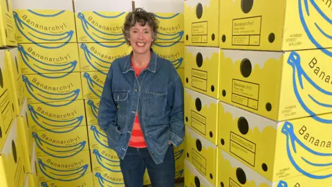 BBC / Sally Fairfax Lady smiling with short wavy hair, blue denim jacket, striped red and orange jumper and blue jeans. She is stood next to a stack of yellow boxes with Bananarchy written on them.