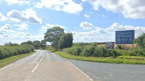 Google A country road flanked by green bushes and trees. There is an exit lane on the right with a big blue sign on the grass verge reading "Welcome to Bridlington". Next to that is a white road sign listing Grindale, Burton Fleming and Malton, with a black arrow pointing right. The sky is blue with scattered white and grey clouds.