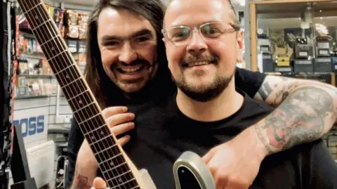 Mark Wood Sam Orr is smiling as he stands behind Mark Wood in Sam's guitar shop. Mark Wood is also smiling as he holds his guitar, with the neck of the guitar and the frets visible.