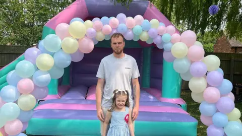 Turner Family James Turner with short dark hair and beard wearing a grey t-shirt stands behind his daughter Emilee who has dark shoulder length hair and is wearing a blue dress and red rosette. They are standing in front of a multi-colour bouncy castle with an arch of balloons