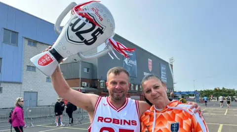 BBC Chris wears a Robins white and red vest and holds up a giant blow up trophy with the Robins 2025 on it. His arm is around his female friend.