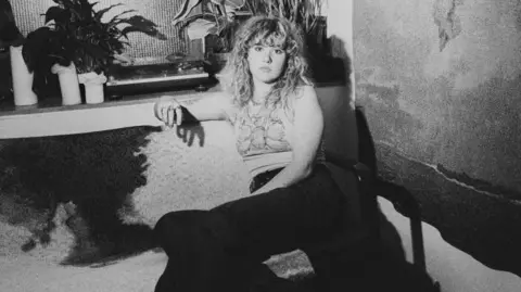 Flower Up A black and white photo of a young woman with curly long hair and a full fringe sat on a sofa and looking into the camera with a serious expression. Her arm is resting on the side of the sofa and you can see plant pots on a shelf behind her. She is wearing a light vest top and dark trousers. 