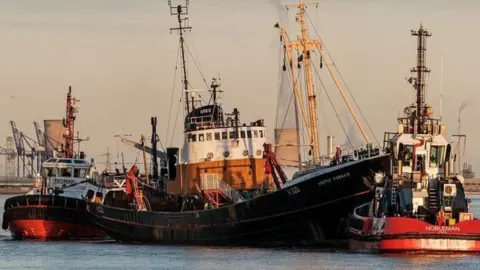 Hull: Yorkshire’s Maritime City The trawler Arctic Corsair being towed on an estuary by two tugboats. It has a black hull with a wood and white steel bridge and a single funnel. The smaller tugs have red hulls and white cabins.