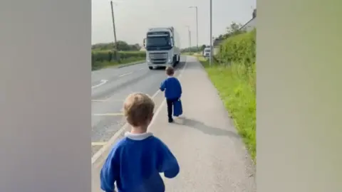 PG Linde Two children in blue school jumpers walking down the road towards the primary school about a metre away from a lorry driving down a main road