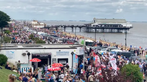 BBC A landscape view of groups of people along the seafront at Cleethorpes. You can see people sat around next to a cafe which has ice creams signs, several cars parked along the promenade and a peer in the distance with a long building at the end.