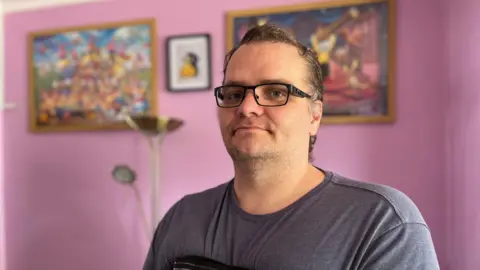 BBC Neil Ferriby. He has short dark hair and wears black rimmed glasses. He is wearing a grey t-shirt and has a black elasticated support bandage around his chest. He is standing in front of a pink wall with three framed prints on it.