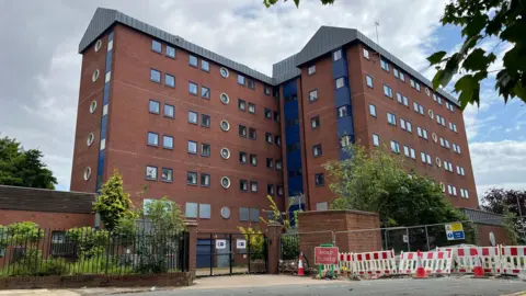 BBC Two large tower blocks made of red bricks, blue cladding and grey panels stand behind a metal fence. The lower doors and windows are covered with metal sheeting. A sign next to the gate reads 'Road closed'.