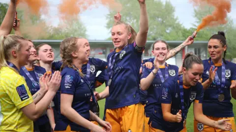 Getty Images Hull City Ladies football team celebrating on the field, wearing navy and amber kits. One player is raising her arm as others are cheering and smiling. Players have blue medals around their necks. Orange smoke flares fill the air. Green trees can be seen in the background.
