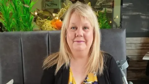 Family handout A woman, in her 50s, with long blonde hair. She is wearing a floral yellow dress under a black cardigan and is smiling 