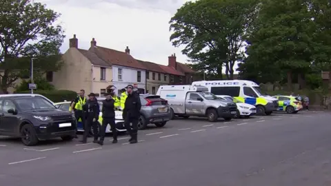 BBC Police officers and police vehicles in a car park in Easington. Officers are wearing dark blue overalls and caps with some wearing high visibility jackets. They are huddled in a group and standing in front of a black Range Rover and a marked police car.