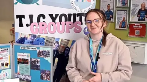 Yasmin Bartle Yasmin Bartle. She has long brown hair and glasses. She is smiling. She has two blue lanyards and a light hoody top. There is a display behind her and next to her with information about Tourette syndrome.