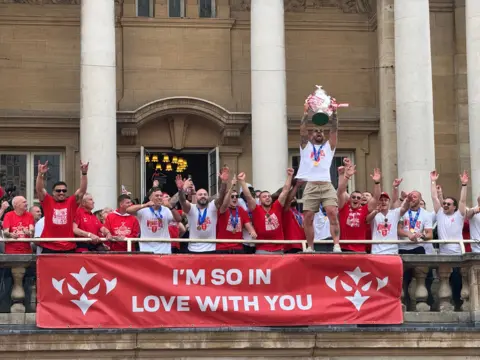 A crowd of rugby league players stand on a balcony one man, wearing a white t-shirt and shorts, is holding a large silver trophy above his head. 