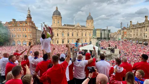 BBC A crowd of rugby league players stand on a balcony overlooking Queen Victoria Square in Hull where thousands of fans, dressed in red and white, have gathered. One player is standing with a large silver trophy above his head. 